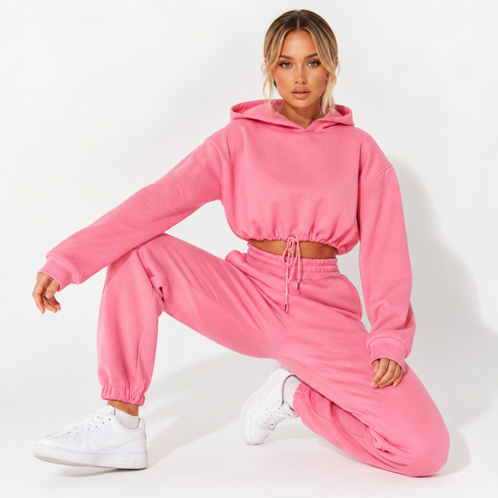 Women tracksuit set in pink on a female model with cropped hoodie, matching joggers and sneakers on a clean white backdrop.